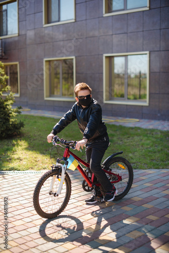 Outdoor activities during quarantine. Boy in a black mask riding a bike in the yard. There is Ukrainian flag on his bicycle. Bad mood about coronavirus