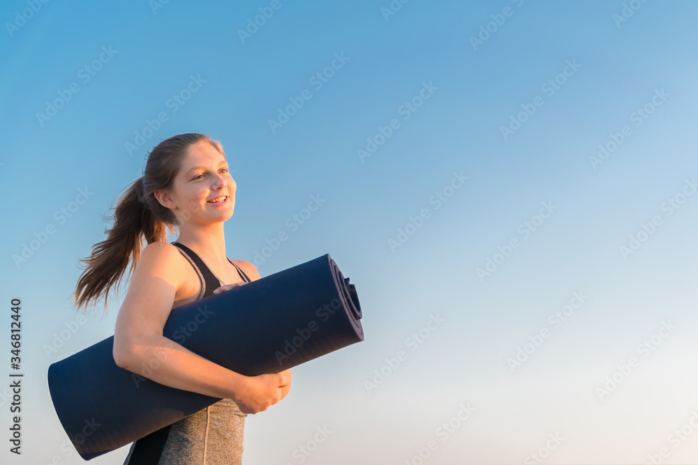 A young millennial smiling girl in sportswear is standing on a blue sky background, holding a rolled mat in her hands A young millennial smiling girl in sportswear is standing on a blue sky background, holding a rolled mat in her hands