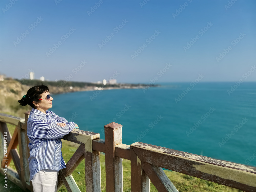 A middle-aged woman in a shirt and sunglasses stands on a high bank above the sea and looking at great distances.