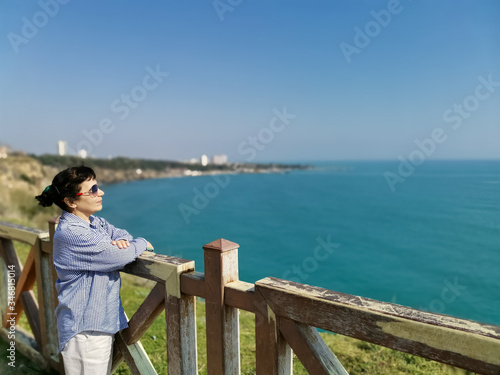 A middle-aged woman in a shirt and sunglasses stands on a high bank above the sea and looking at great distances. © Galina