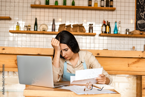 Upset cafe owner holding paper near calculator, laptop and documents with glasses at table