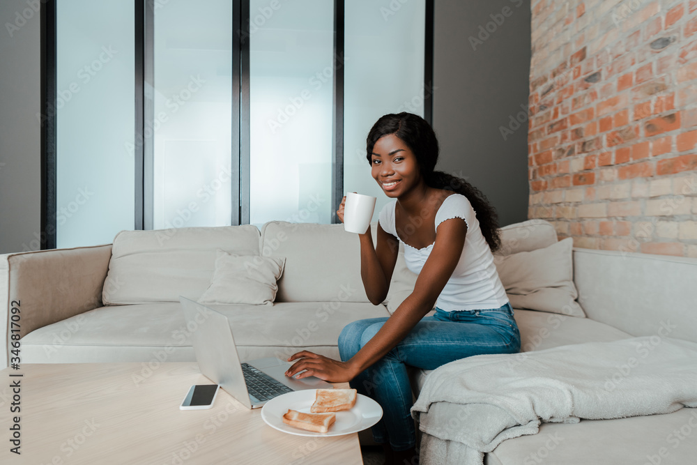 African american freelancer with cup of tea smiling and using laptop at coffee table with smartphone and plate with toasts in living room African american freelancer with cup of tea smiling and using laptop at coffee table with smartphone and plate with toasts in living room