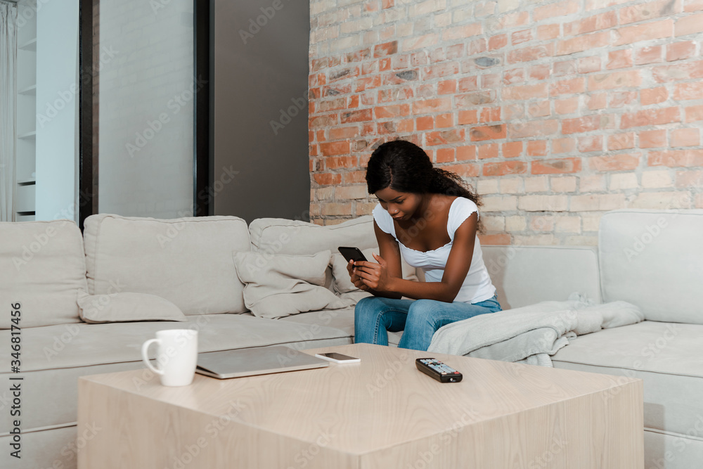 African american woman with digital tablet on sofa near coffee table with laptop, smartphone, remote controller and cup of tea in living room African american woman with digital tablet on sofa near coffee table with laptop, smartphone, remote controller and cup of tea in living room