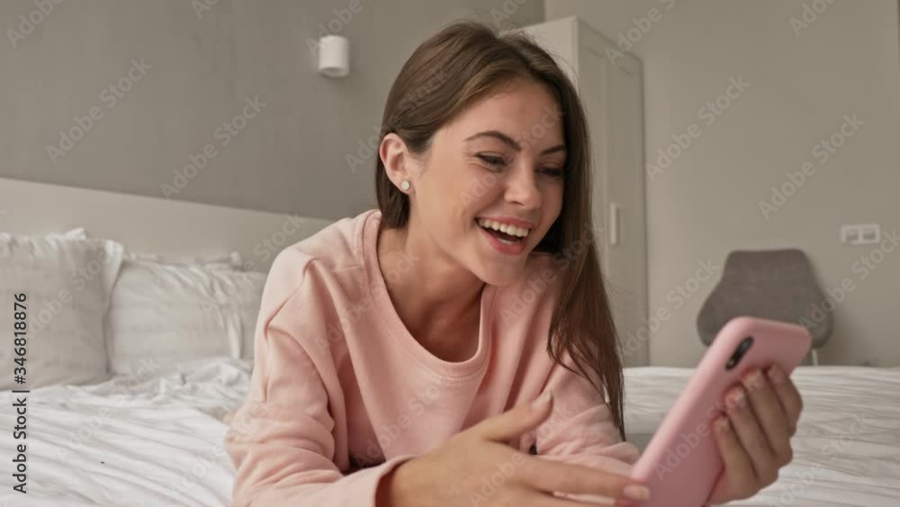 A happy young attractive girl is having a video call using her smartphone lying in the bed and at home A happy young attractive girl is having a video call using her smartphone lying in the bed and at home