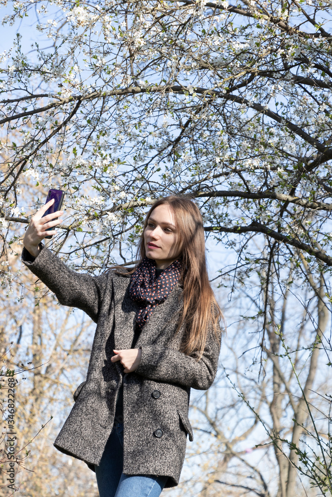 Fashionable girl in spring park makes selfie. Dressed in a coat and jeans, with sunglasses. Beauty. Fashionable girl in spring park makes selfie. Dressed in a coat and jeans, with sunglasses. Beauty.
