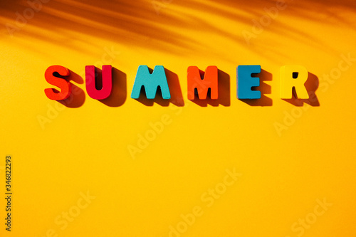 Flat lay composition with phrase SUMMER made of colorful wooden letters on yellow background, with palm leaf shadow next to it, and available copy space.