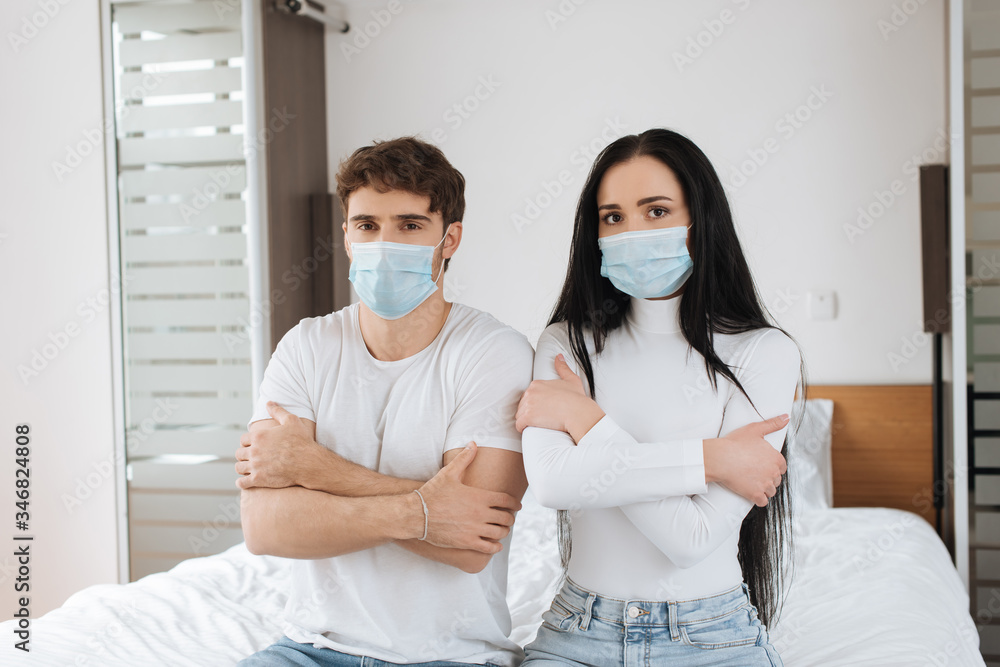 cold couple in medical masks sitting on bed during self isolation