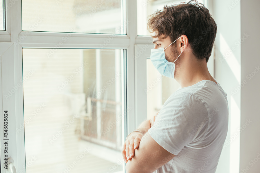 sad ill man in medical mask with crossed arms looking through window during self isolation sad ill man in medical mask with crossed arms looking through window during self isolation