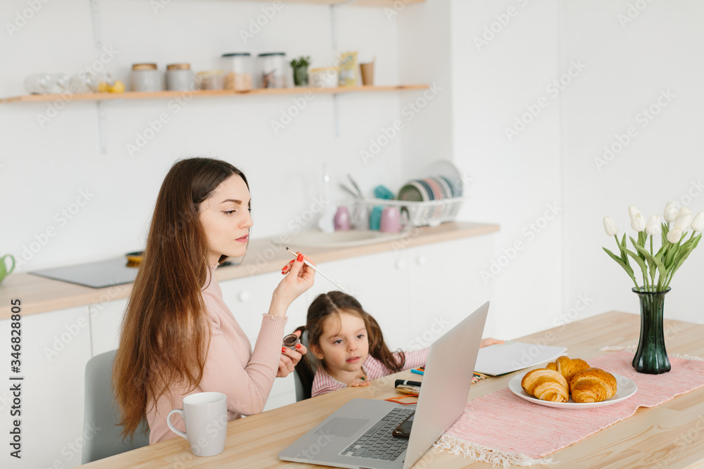 Smiling pretty woman doing make up while sitting with her little daughter at the kitchen during breakfast.