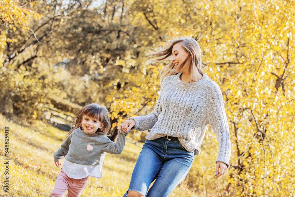 Fototapeta premium Beautiful woman and her little daughter holding hands and running in autumn park. Tiny girl having fun and playing with her young mother outdoors. Happy family relationships of mom and child