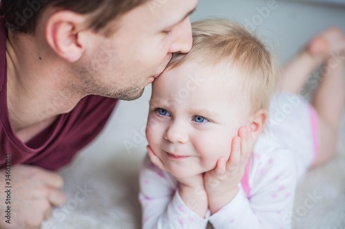Blond-haired blue-eyed baby girl and young dad in the home interior. Dad kisses his little daughter.