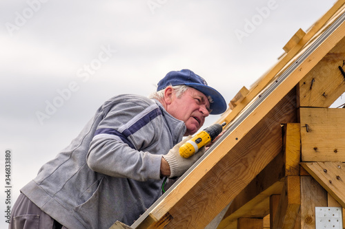 Senior gray-haired Construction man using a screwdriver, fastens a roofing sheet to wooden rafters on the roof of a country house under construction. The physical activity of the elderly.