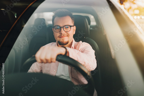 A young europen beautiful smiling man wearing glasses driving a car in the sity on sunset. View through the windshield