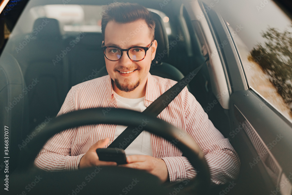 Obraz premium A car driver sitting in a car with seat belt and smartphone. View .through the windshield of a stylish smiling man in glasses