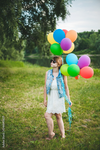 Beautiful cheerful brown-haired girl in sunglasses stands and smiles while holding multi-colored balloons. The girl cheerfully puffed out her cheeks.