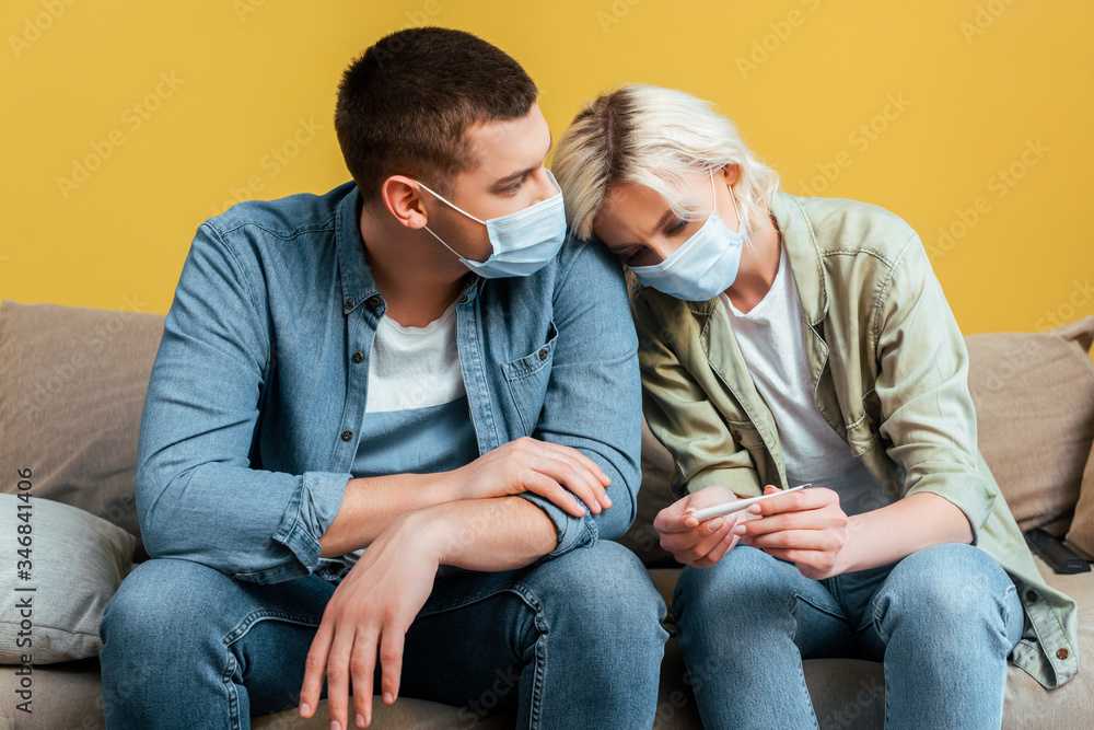 sad young couple in medical masks with thermometer on sofa near yellow wall