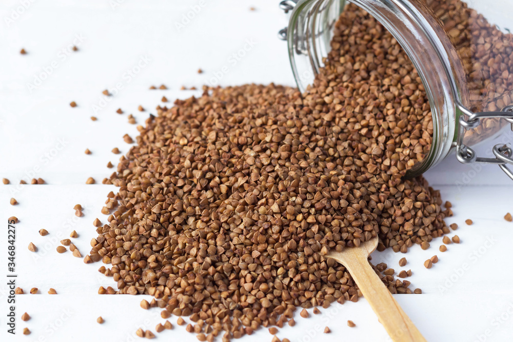 buckwheat is scattered on a table from a glass jar, next to a spoon buckwheat is scattered on a table from a glass jar, next to a spoon