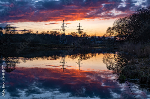 Two towers of a power line against the background of an orange sunset and their reflection in the water. © Valery Smirnov
