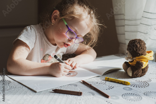 A little girl with glasses draws a portrait of a soft teddy bear, which lies in front of her. Idea for quarantining a child