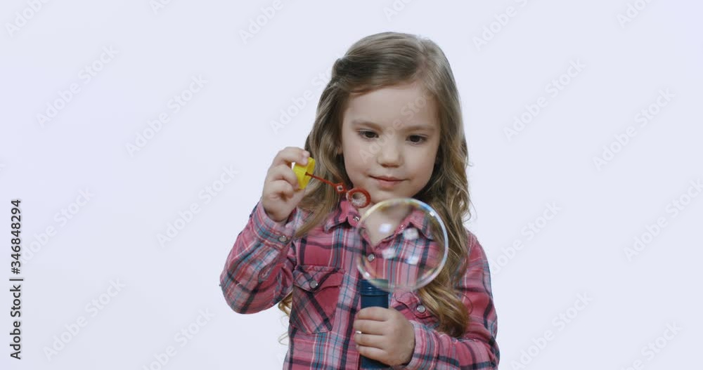 Cute Caucasian blonde and joyful little girl with fair hair blowing soap bubbles and feeling happy on the white wall background. Cute Caucasian blonde and joyful little girl with fair hair blowing soap bubbles and feeling happy on the white wall background.