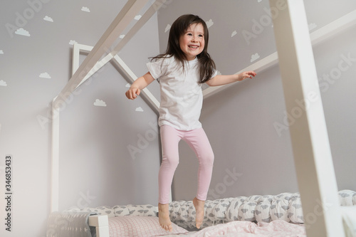 Happy little girl wearing white pajamas jumping on bed in white bedroom. Cute girl having fun while jump and play