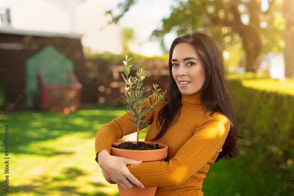 Beautiful dark haired smiling woman holding pot with plant while standing in the garden