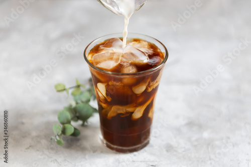 Milk is pouring into a tall glass with brew black coffee with pieces of ice on a gray dark background, close-up. Ice summer fresh coffee concept