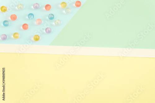 Bright color block paper background (yellow, light blue and mint green) with some multicolored pastel beads and white ribbon. Copy space. Space for text. Flat lay, top view.