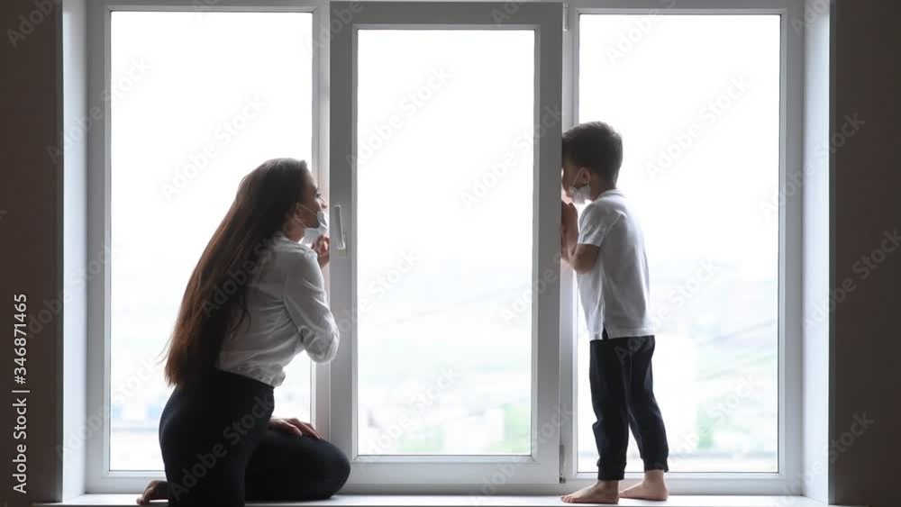 A woman with a child in medical masks is quarantined at home and playing near the window during the worldwide pandemic of the coronavirus and Covid -19 A woman with a child in medical masks is quarantined at home and playing near the window during the worldwide pandemic of the coronavirus and Covid -19