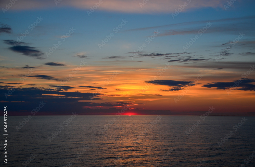 Sun is setting behind the Baltic Sea. The sky is colorful combination of yellows, oranges and blues. The photo is a dramatic because of the big clouds, and sea with waves. Very scenic.