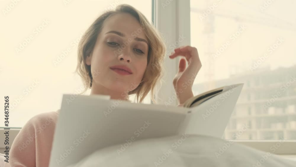 Happy blonde optimistic woman indoors at home in bed reading book Happy blonde optimistic woman indoors at home in bed reading book