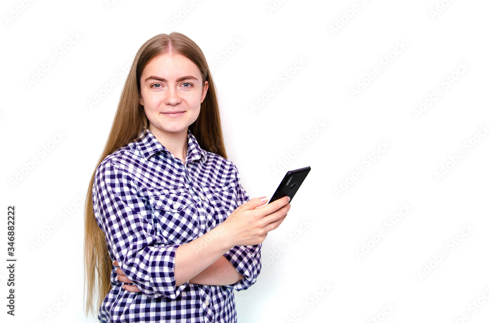 A young girl in a plaid shirt with long hair stands on a white background and holds a smartphone in his hand. A girl looks, is surprised, scared, rejoices looking at a smartphone A young girl in a plaid shirt with long hair stands on a white background and holds a smartphone in his hand. A girl looks, is surprised, scared, rejoices looking at a smartphone
