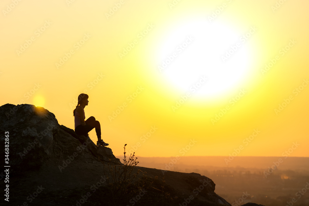 Fototapeta premium Silhouette of a woman hiker sitting alone on big stone at sunset in mountains. Female tourist on high rock in evening nature. Tourism, traveling and healthy lifestyle concept.