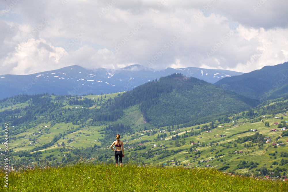 Back view of slim young woman standing on grassy valley on background of green mountains on sunny summer day.
