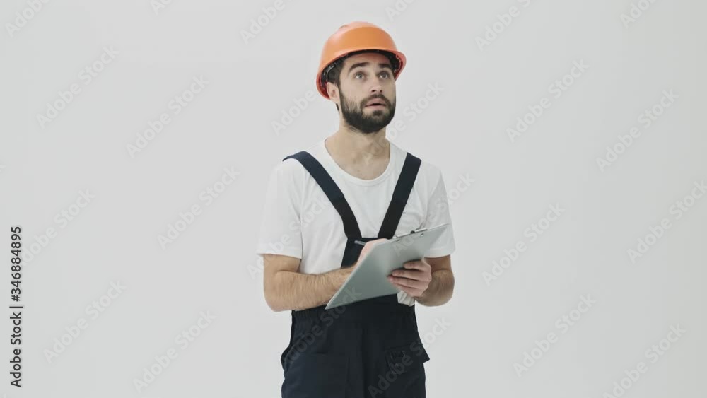 Concentrated young bearded man builder isolated over white wall background in helmet writing notes in clipboard. Concentrated young bearded man builder isolated over white wall background in helmet writing notes in clipboard.