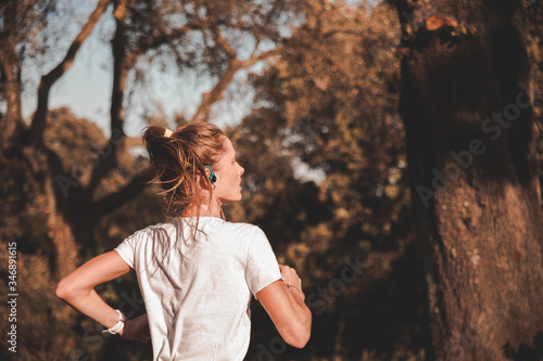 Giovane ragazza runner si allena da sola e concentrata indossando auricolari in mezzo alla natura di un parco urbano 
