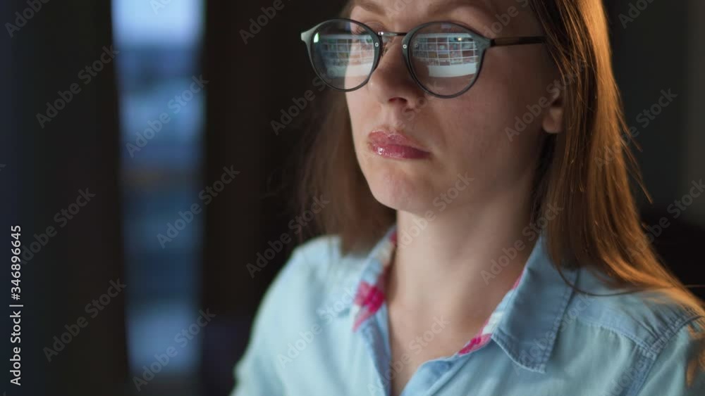 Woman in glasses looking on the monitor and surfing Internet. The monitor screen is reflected in the glasses. Work at night. Home Office. Remote work Woman in glasses looking on the monitor and surfing Internet. The monitor screen is reflected in the glasses. Work at night. Home Office. Remote work