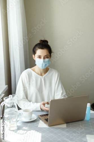 woman in medical mask, hand sanitizer on the table, working with a laptop notebook, online learning education shopping