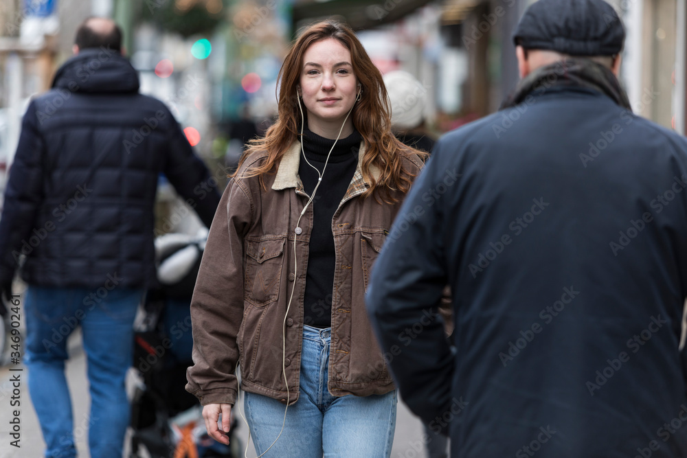 Fototapeta premium Young Woman Wearing Jeans and Brown Coat Walking Amongst Pedestrians