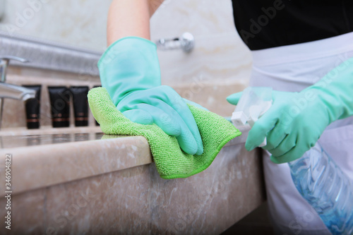 Women's hands in green rubber gloves clean the edge of the marble countertop in the bathroom. An unrecognizable photo. The concept of cleanliness and hygiene in a hotel or at home.