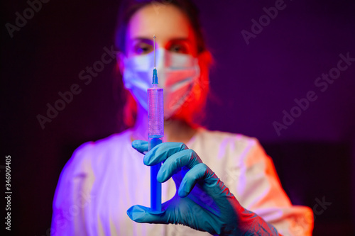 Medical worker in a medical mask with a syringe in hands. young medic girl with a syringe in a dark room with red and blue light. medical worker during the coronavrius pandemic photo