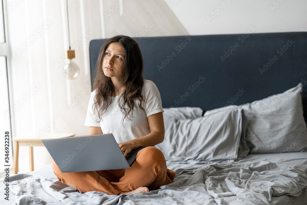 Beautiful young brunette girl with a laptop sitting on the bed. Stylish modern interior. A cozy workplace. Shopping on the Internet Beautiful young brunette girl with a laptop sitting on the bed. Stylish modern interior. A cozy workplace. Shopping on the Internet