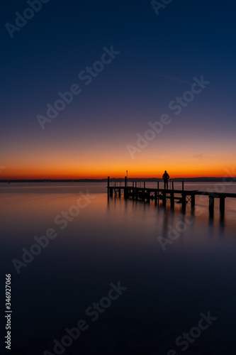 Fototapeta Naklejka Na Ścianę i Meble -  A man silhouette standing on wooden pier lonely at the sea with beautiful sunset. lsunset seascape at a wooden jetty.