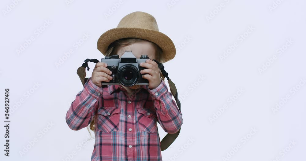 Portrait shot of the cute little Caucasian girl in a hat smiling joyfully to the camera and holding photocamera, then taking a photo. White background. Portrait shot of the cute little Caucasian girl in a hat smiling joyfully to the camera and holding photocamera, then taking a photo. White background.