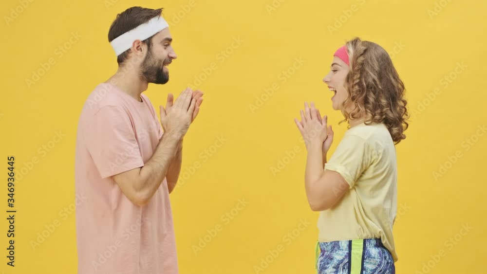 A happy cheerful young athletic couple man and woman in sportswear are playing pat-a-cake while isolated over a yellow background in studio A happy cheerful young athletic couple man and woman in sportswear are playing pat-a-cake while isolated over a yellow background in studio