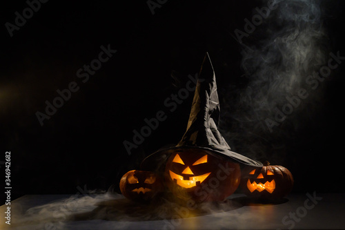 Spooky orange pumpkins for Halloween stand in a row on a dark background. Jack O Lantern in a witch hat. Mystical fog creeps on the ground. Trick or treat.
