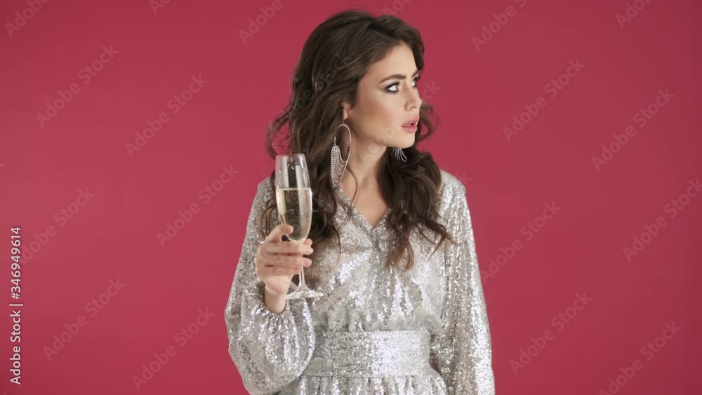 A sad young woman in bright sequins dress is drinking champagne isolated over pink wall background A sad young woman in bright sequins dress is drinking champagne isolated over pink wall background