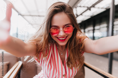 Good-humoured female model with straight hair making selfie with smile. Indoor shot of laughing ecstatic woman in pink sunglasses.