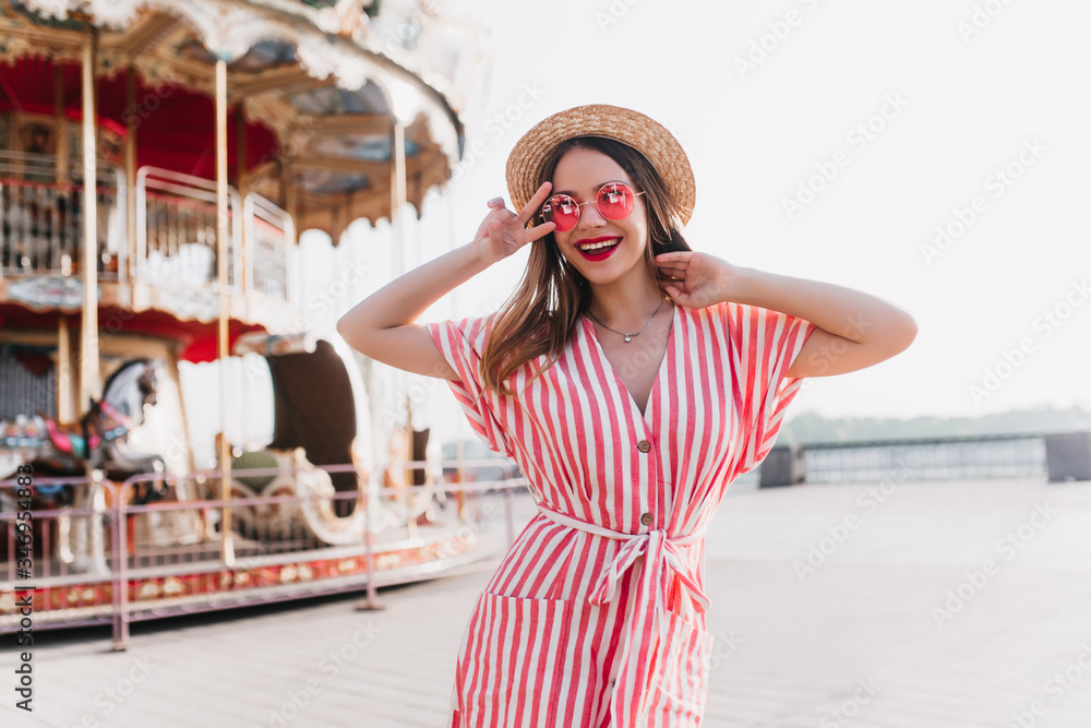 Enthusiastic blonde girl in stylish sunglasses enjoying event in amusement park. Outdoor photo of glad slim woman in straw hat and striped dress standing beside carousel.
