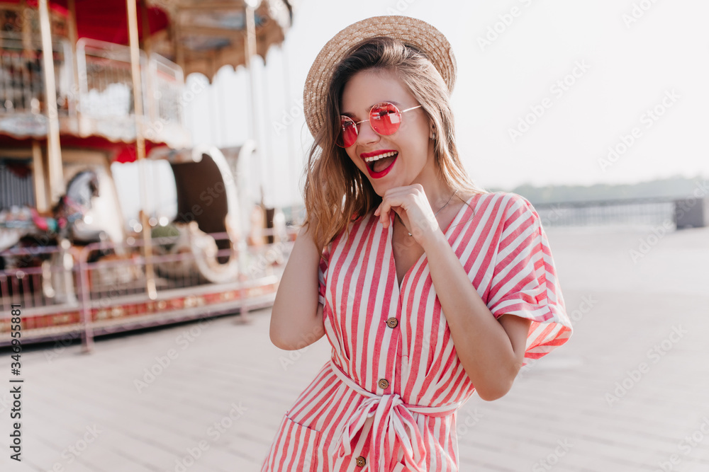 Fototapeta premium Lovely white girl in straw hat laughing while posing beside carousel. Stunning blonde young lady spending summer day in amusement park.